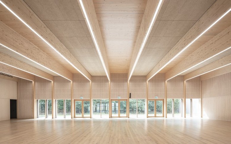 Im Innen der Kulturhalle dominieren helle Sichtholzflächen, der Blick schweift durch die fast gebäudebreite Glasfront in den direkt angrenzenden Bienwald. Bild: AV1 Architekten GmbH, Kaiserslautern
